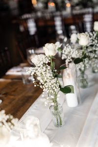 A close-up of a table decorated with white roses and baby's breath in glass vases, alongside a white candle in a glass holder on a white runner, captures an elegant wedding editorial scene perfect for any photographer seeking inspiration.