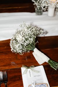 A bouquet of white baby's breath flowers rests on a wooden table next to a folded menu tied with a ribbon, with a fork and knife placed nearby—an elegant setting perfect for wedding photography in Toronto.