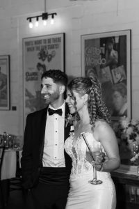 A smiling bride and groom stand together indoors, the bride holding a glass. Both wear formal attire, captured beautifully by the photographer. Framed posters and flowers are visible in the background, adding depth to this editorial-style photography moment.
