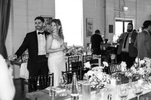A smiling couple, dressed in wedding attire, stands together among decorated tables at a formal event. Captured with a documentary flair, guests mingle in the background. The scene is lively and elegant.