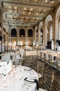 Elegant wedding reception in a grand hall with high ornate ceilings, large arched windows, round tables with white linens, gold chairs, candles, and white floral centerpieces—perfect details for any photographer to capture stunning wedding photography.