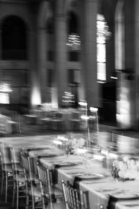 Black and white photo of an elegant, blurred editorial wedding dining setup in a large hall with chandeliers, decorated tables, and rows of chairs.