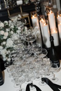 A pyramid of empty champagne glasses is arranged onto a table, with bottles of Prosecco nearby. Lit candles and white flower arrangements lend an editorial photography feel to the background, creating an elegant atmosphere.