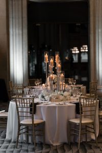 A round table with a white tablecloth is set for six, featuring elegant glassware, white flowers, and a tall centerpiece with lit candles. Gold chairs surround the table in a dimly lit, formal setting, creating an editorial atmosphere perfect for any photographer seeking sophisticated inspiration.