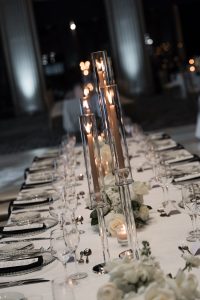 Elegant table setting with tall glass candle holders, white flowers, plates, cutlery, and wine glasses arranged for a formal dinner in a softly lit, upscale venue—perfect for an editorial photography shoot or to capture by any discerning photographer.