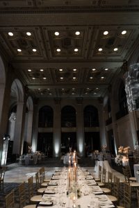 Elegant banquet hall with high ornate ceiling, tall arches, gold chairs, and a long dining table set for a wedding with candles, plates, and glasses. Soft lighting creates a sophisticated atmosphere perfect for an editorial celebration or formal event.