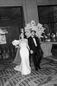 A bride in a strapless gown and veil holds a bouquet and smiles while holding hands with a groom in a tuxedo as they walk together indoors, surrounded by elegant decor and large floral arrangements, while a photographer captures every special wedding moment.