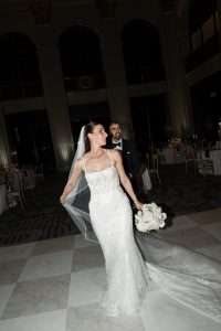 A bride in a white gown and veil holds a bouquet and smiles, walking indoors with a groom in a tuxedo behind her. Capturing an editorial photography style, the elegant room features tall columns and round tables.