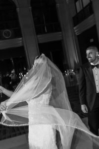 A bride has her veil lifted while smiling and dancing with a man in a tuxedo at a formal indoor wedding. A photographer captures the joyful moment, which takes place in an elegant, dimly lit setting.