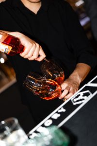 A bartender in black pours rosé or a pink cocktail into two large wine glasses at a bar, with part of a drink and bar mat visible in the foreground, capturing a documentary-style moment as if seen through the lens of a photographer at a wedding.