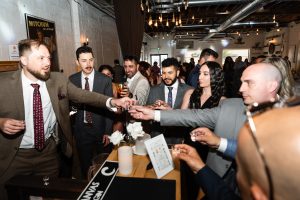 A group of people in formal attire stand around a bar, raising shot glasses together for a toast in a well-lit, modern indoor venue. White flowers and a candle are on the counter, perfectly capturing the joyful spirit of the wedding as documented by the photographer.