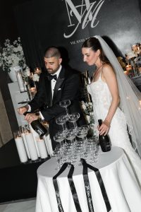 A bride and groom in formal wedding attire pour champagne onto a pyramid of glasses at a decorated reception, with candles and flowers in the background, capturing an elegant moment of editorial photography.
