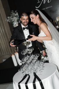 A bride and groom in formal attire pour champagne together onto a tower of glasses at their wedding reception, smiling and celebrating. A wedding photographer captures the moment as white flowers and candles decorate the background.