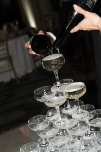 A photographer captures the moment as a person pours champagne from a bottle onto the top glass of a tiered champagne tower, causing the sparkling wine to overflow into the glasses below.