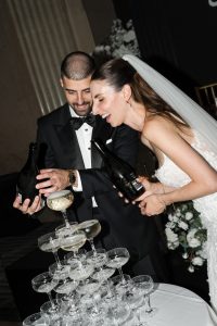 A bride and groom in formal wedding attire happily pour champagne together onto a tower of glasses, with white flowers and elegant decor creating an editorial-worthy background.