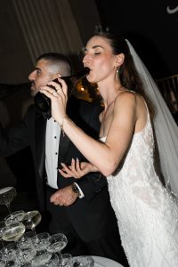 An editorial-style image captures a bride in a white dress and a groom in a tuxedo drinking from dark bottles at a wedding celebration, standing beside a table with stacked champagne glasses—a perfect moment for any wedding photographer to remember.