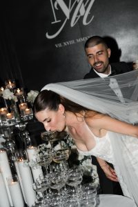 A bride in a white dress and veil drinks from a champagne glass at the top of a tower, while a smiling groom stands behind her. Candles and flowers decorate the background, capturing an elegant wedding moment that any photographer would cherish for their editorial portfolio.
