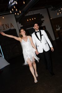A smiling couple, holding hands, walks indoors in true editorial style. The woman wears a fringed white dress and heels, while the man wears a white tuxedo jacket and black pants. They appear joyful, exuding wedding excitement as the woman waves her arm—capturing an unforgettable moment in Toronto.