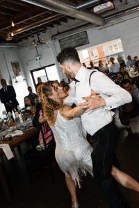 A couple in formal attire dances energetically at a lively indoor wedding, with guests seated at decorated tables in the background watching and smiling, captured beautifully by a documentary photographer.