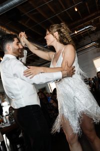 A couple dressed formally dances joyfully together indoors, with the woman in a white, fringed dress and the man in a white jacket. Exposed pipes and wooden beams are visible on the ceiling above them, adding an editorial flair to this photography scene.