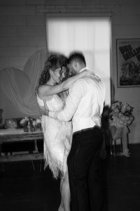 A couple dressed in formal attire shares a close dance in a dimly lit room, with a heart-shaped decoration and flowers visible in the background, capturing a romantic wedding moment reminiscent of an editorial photoshoot in Toronto.