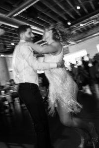 A man and woman joyfully dancing together indoors, captured in motion blur by a talented Toronto photographer. The woman wears a sparkly dress; the man is in a white shirt. Other people and tables are visible in the background, adding to the lively, editorial feel of the scene.