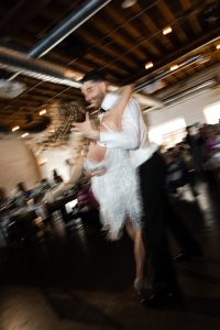 A couple dances energetically in a blurred, motion-filled photo at an indoor event with exposed ceilings and seated guests in the background, capturing the spirit of documentary photography in Toronto.