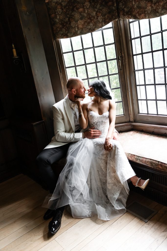 A bride and groom sit by a window in a cozy, rustic room, gazing lovingly at each other in this Toronto wedding editorial. The bride wears a lace wedding dress, and the groom is in a light-colored suit. Natural light streams through the window.