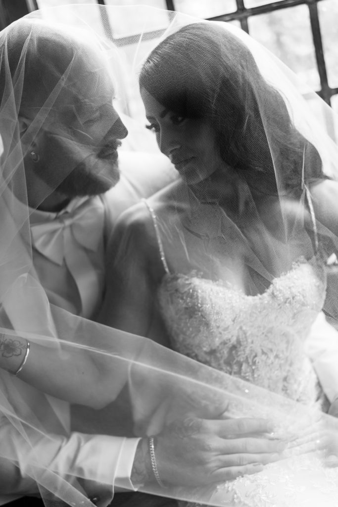 A bride and groom sit closely together under a sheer veil, gazing at each other lovingly. Captured by a talented Toronto photographer, the black and white image has an editorial quality, highlighting an intimate moment on their wedding day.