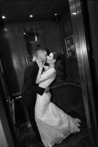 A bride and groom share a romantic kiss in an elevator, dressed in wedding attire. The black and white photo, taken by a skilled wedding photographer, captures an intimate moment on their special day.