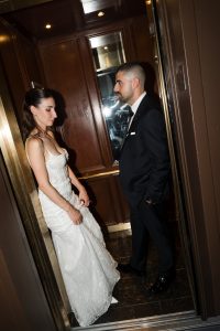 A woman in a white wedding dress and a man in a dark suit stand facing each other inside an open elevator with wood paneling, sharing a quiet, editorial moment together.