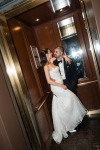 A bride and groom in formal wedding attire embrace and smile at each other inside a wood-paneled elevator, as a photographer captures the moment—the camera flash reflecting onto the mirror behind them.