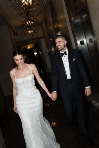A bride in a white gown and a groom in a dark tuxedo hold hands and smile while walking in a hallway with elevators and modern light fixtures, beautifully captured by the photographer.