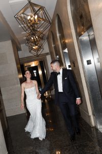 A bride in a white dress and a groom in a black tuxedo walk hand in hand, smiling, onto a modern hallway with geometric light fixtures and elevator doors, capturing an elegant wedding editorial moment.