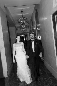 A couple dressed in wedding attire, holding hands and smiling, walk down a hallway with geometric light fixtures and tiled floors. The black-and-white photography lends an editorial elegance to the image.