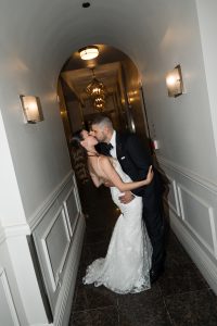 A couple in formal wedding attire shares a kiss in a narrow, elegant hallway with arched ceilings and wall sconces, capturing an editorial moment of romance through stunning wedding photography.