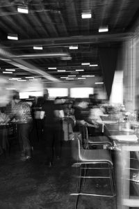 Black-and-white photo by a Toronto photographer captures a blurred, busy restaurant or cafe scene with people moving around tables and chairs, suggesting motion and activity. This documentary-style image is taken indoors with industrial-style ceilings and lighting.