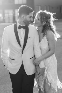 A couple dressed formally, smiling at each other while holding hands. Captured with a touch of documentary photography, the moment feels candid and genuine—the man in a white tuxedo jacket and bow tie, the woman in a sleeveless, sparkly dress. They are outdoors in soft sunlight.