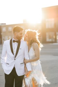 A couple dressed formally stands outdoors at sunset, captured in beautiful wedding photography. The woman in a white dress embraces the man in a white tuxedo jacket and black bow tie. Sunlight glows behind them, creating a warm, romantic atmosphere reminiscent of a timeless documentary.