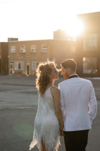 A couple dressed in formal attire holds hands and walks outdoors at sunset, with sunlight illuminating their faces and an urban background behind them, capturing a timeless wedding photography moment.