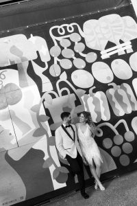 A couple dressed in formal attire smiles at each other while standing in front of a colorful abstract mural. Captured through the lens of skilled documentary photography, the photo is rendered in striking black and white.