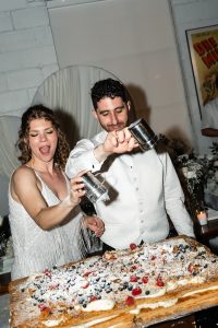 A smiling couple in white outfits joyfully sprinkle powdered sugar on a large dessert topped with berries at a celebratory event, while a wedding photographer captures the delightful moment.