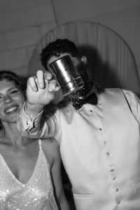 A smiling couple in formal attire stands together, evoking the charm of a classic wedding portrait. The person on the right holds a metal cup toward the camera, sprinkling powder or glitter that partially obscures their face. This black and white image captures a candid, documentary feel, perfect for commemorating a Toronto celebration.