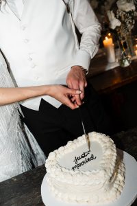 A couple, dressed in formal wedding attire, cuts a heart-shaped cake with Just married written on it. Their hands are together on the knife, capturing a perfect documentary moment of their Toronto wedding, while lit candles and flowers glow softly in the background.