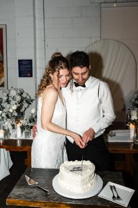 A bride and groom in formal attire cut their wedding cake together, smiling. Captured in a documentary style, the moment is set amidst white flowers and candles decorating the wooden table behind them at their Toronto wedding.