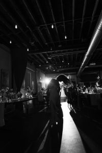 A couple dances closely in a dimly lit room, backlit by bright light. With a documentary style of photography, the moment captures guests seated at decorated tables in the background, creating dramatic shadows and a romantic atmosphere—perfect for an unforgettable Toronto evening.