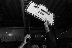 A person holds up a lighted sign reading Just Married! S & M 06.06.26 at a celebration in Toronto, with people and industrial decor visible in the background, perfectly captured by an editorial photographer.