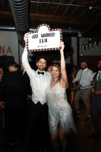 A joyous couple celebrates, holding a light-up sign reading “JUST MARRIED! S & M 06.06.26” at a lively indoor wedding reception with guests dancing in the background, capturing a perfect moment of documentary photography.