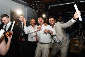 A group of men in suits laugh and pose energetically at a lively indoor party, holding rolled papers and drinks, with warm lighting and people mingling in the background—captured in a candid moment of editorial photography reminiscent of an elegant wedding celebration.