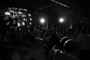 A group of people dance and embrace each other in a dimly lit room, illuminated by bright spotlights, creating dramatic shadows and a festive atmosphere—capturing a powerful moment of connection that any documentary photographer would seek to immortalize through photography.